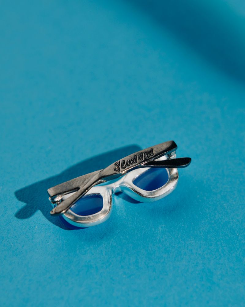 Colgante unisex con forma gafas cuya montura es de plata y las patillas están chapadas en oro negro. Las lentes son la piedra calcedonia azul. En sus bordes tiene una circonita blanca
