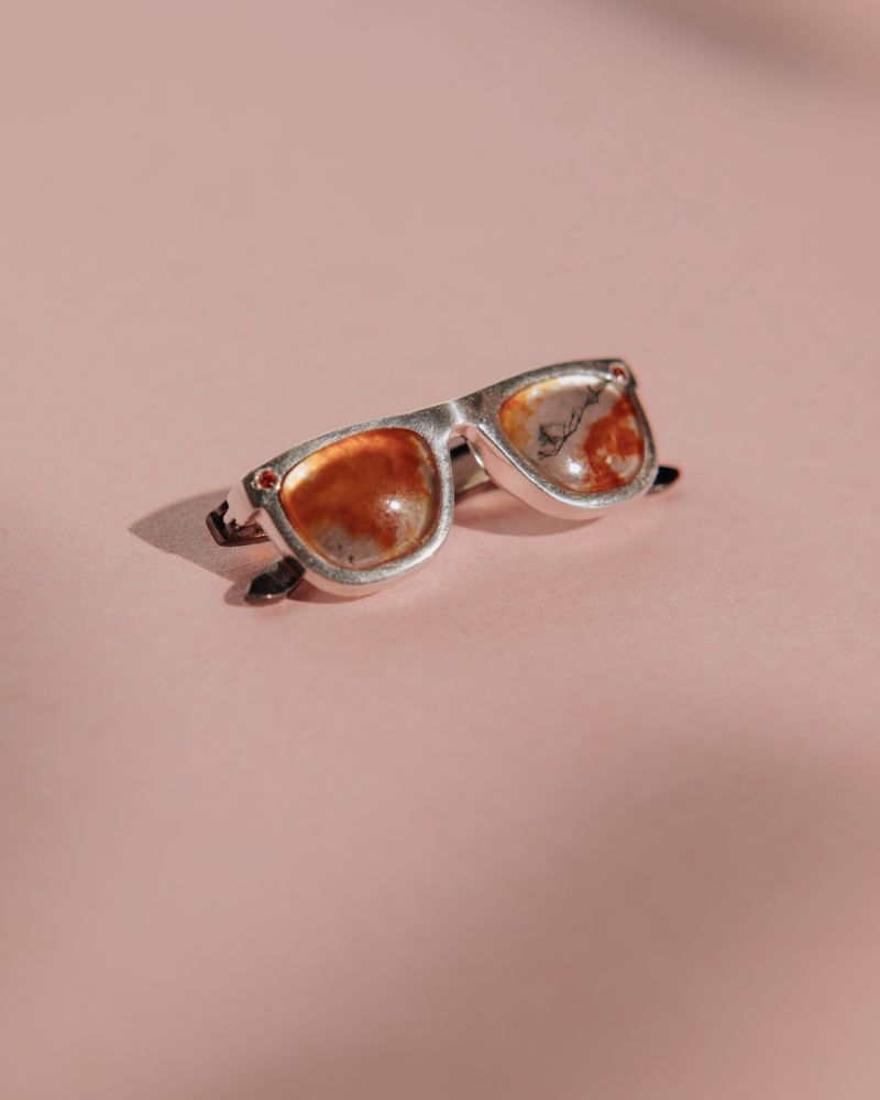 Colgante de gafas unisex con la montura de plata y chapada en oro negro. Los cristales son un cuarzo rojo con dendrítas. Sus circonitas naranjas en los laterales y las patillas chapadas en oro negro recuerdan a las gafas de sol Rayban