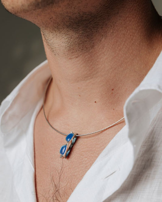 Colgante Unisex con forma gafas cuya montura es de plata y las patillas están chapadas en oro negro. Las lentes son la piedra calcedonia azul.