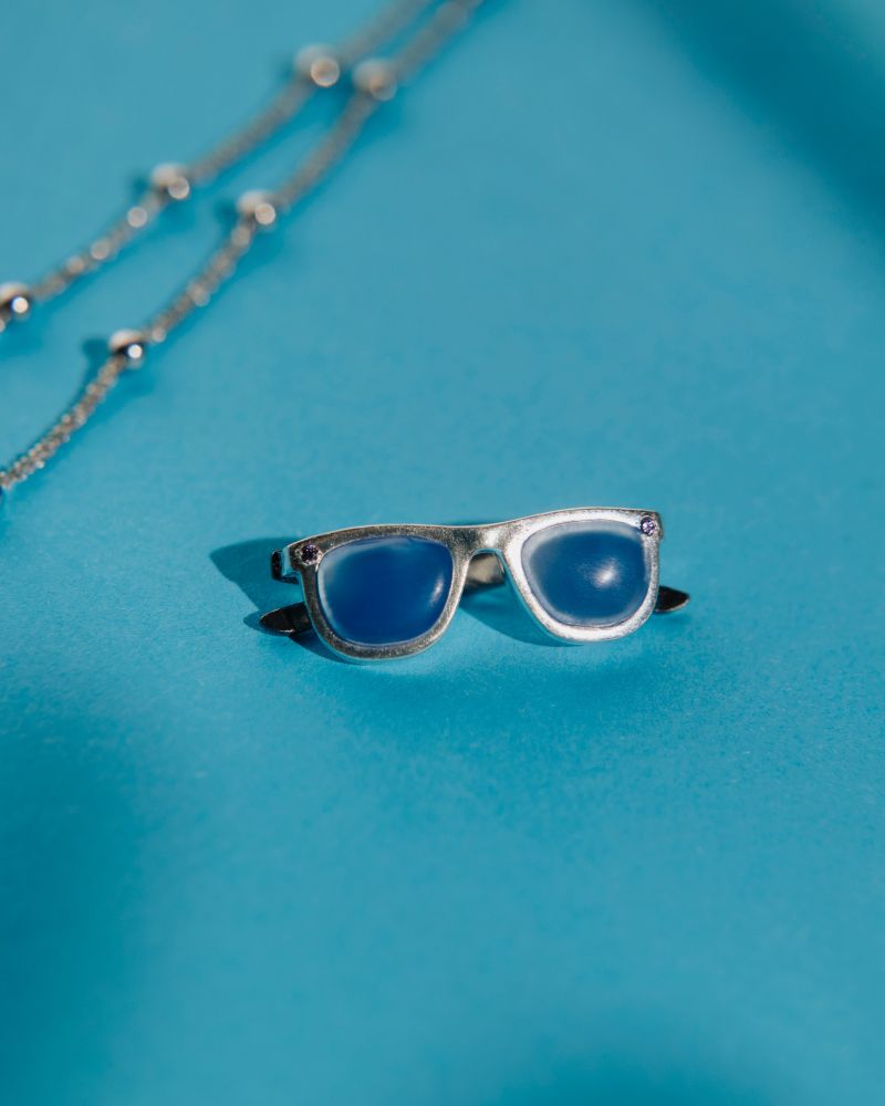 Colgante con forma gafas cuya montura es de plata y las patillas están chapadas en oro negro. Las lentes son la piedra calcedonia azul.